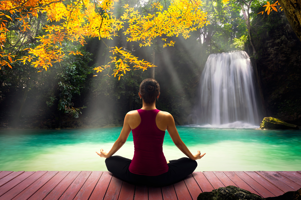 A woman meditating on a dock by a beautiful waterfall flowing into green water.